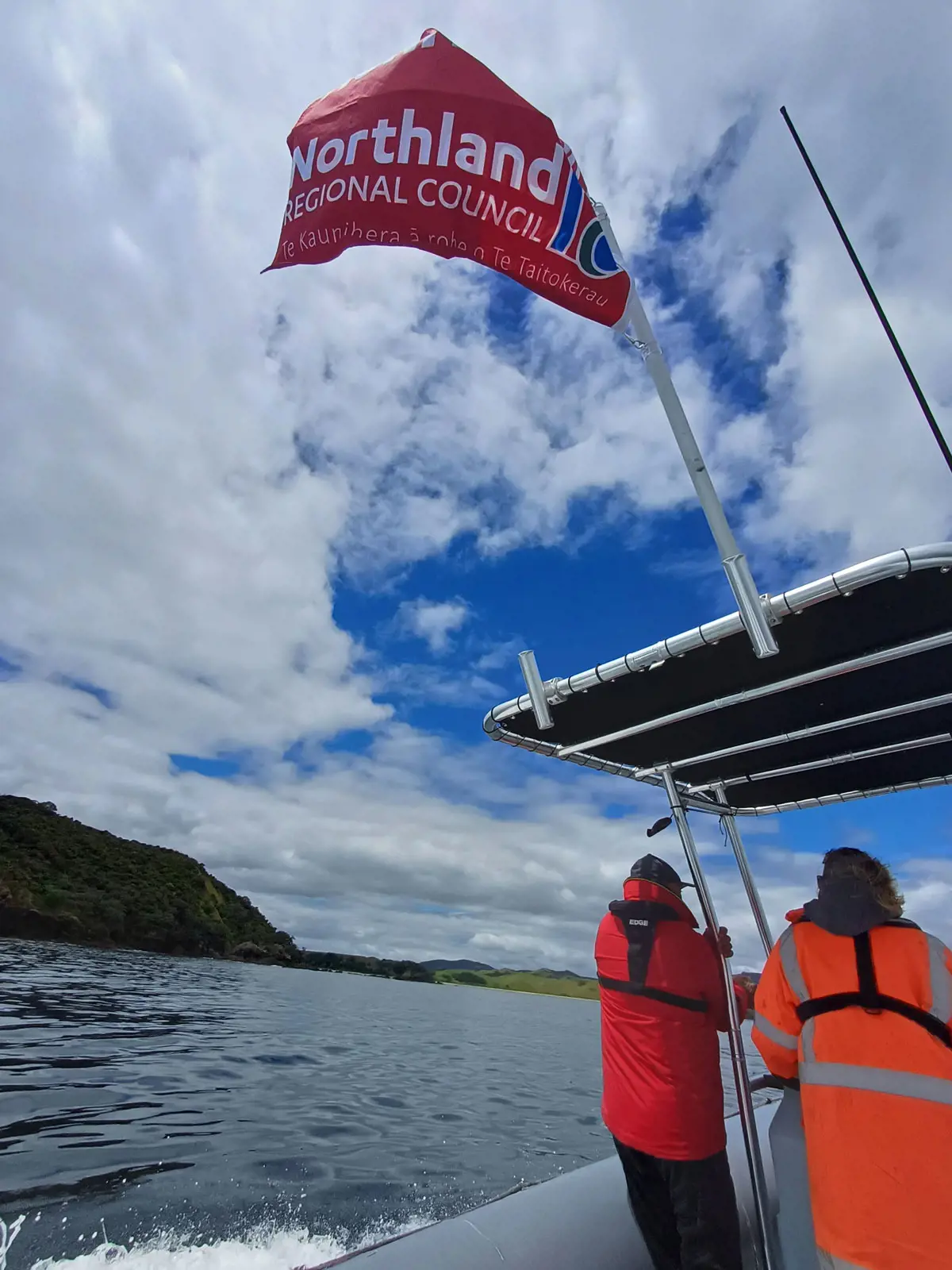 Northland Regional Council staff aboard a patrol vessel on a harbour, backs to the camera, NRc flag visible above