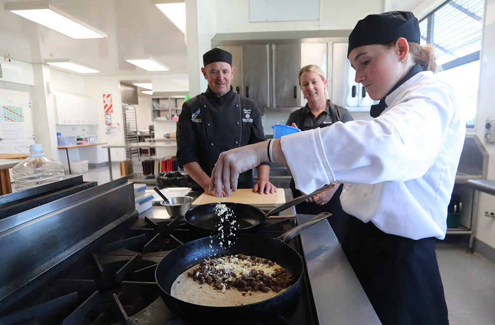 Chef student sprinkles cheese on food while two people watch on.