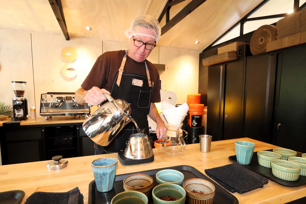 Man wearing hairnet pours water into coffeepot, espresso machine in background